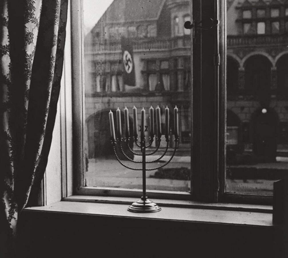 A historic black-and-white photo of a menorah placed in a window across from a Nazi party building, with a Nazi flag visible.